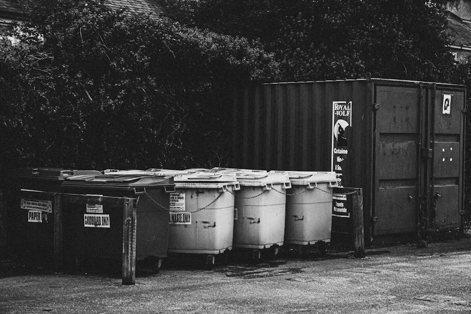 Photograph of an overflowing waste disposal area in an urban setting, featuring a large grey mixed paper and cardboard recycling bin in the center, filled to capacity with torn and crumpled paper, fla