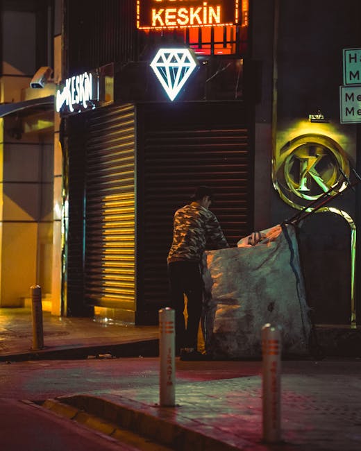 A row of dark green refuse bags, some with visible yellow drawstrings, lined up on a paved sidewalk in front of a retail store window. The bags are filled with various discarded items and are placed d