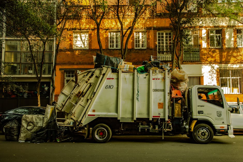 A large collection of mixed waste and rubbish stacked outside a commercial building on a paved street, including black, green, and red bins filled with various discarded items such as cardboard boxes,